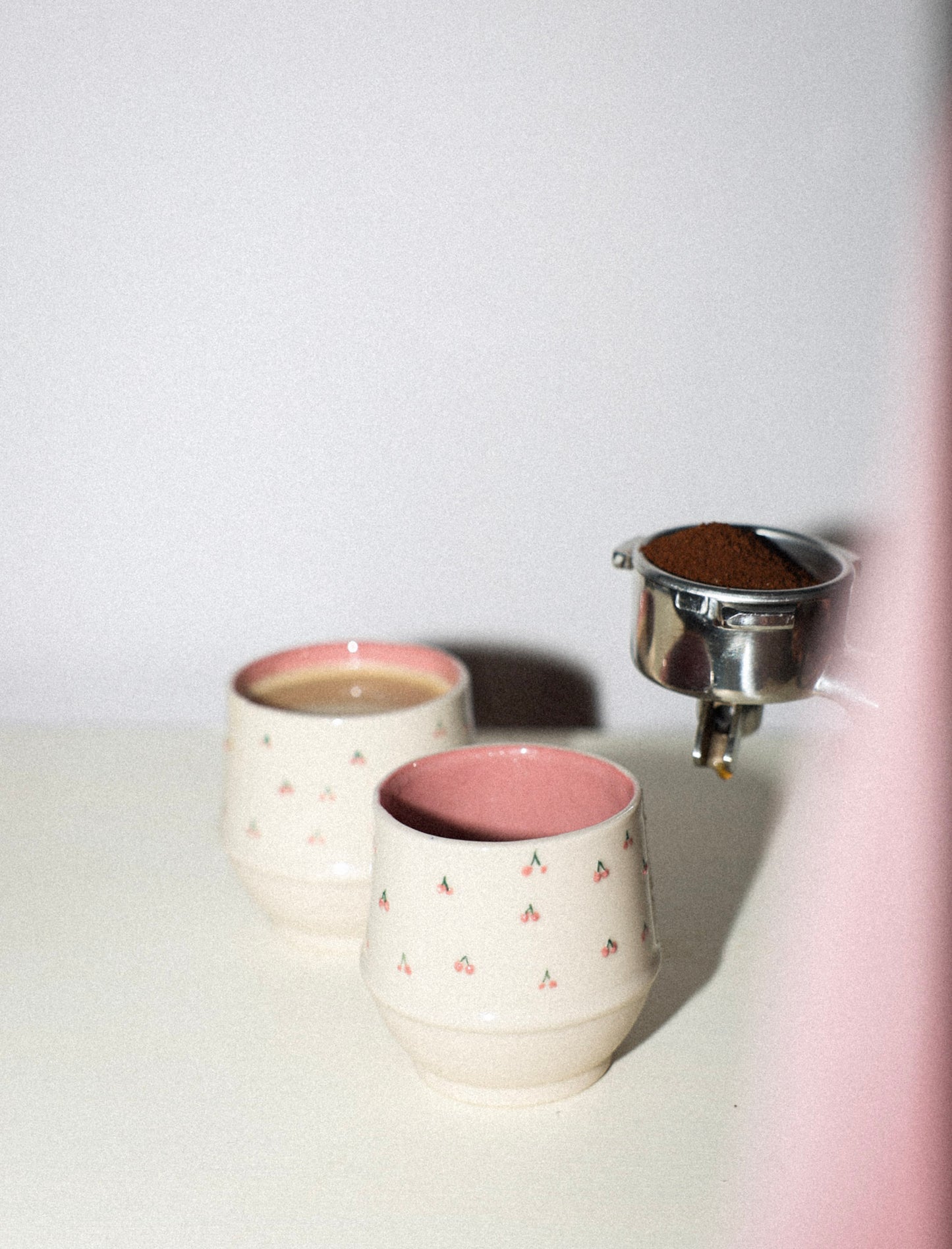 Two ceramic cups with pink interior and exterior, one being filled with a brown substance from a metal container.