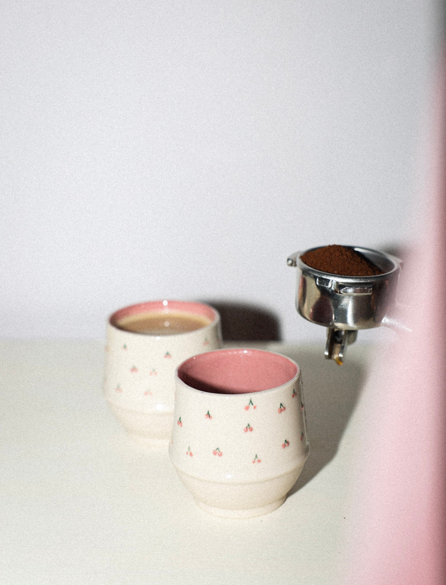 Two ceramic cups with pink interior and exterior, one being filled with a brown substance from a metal container.