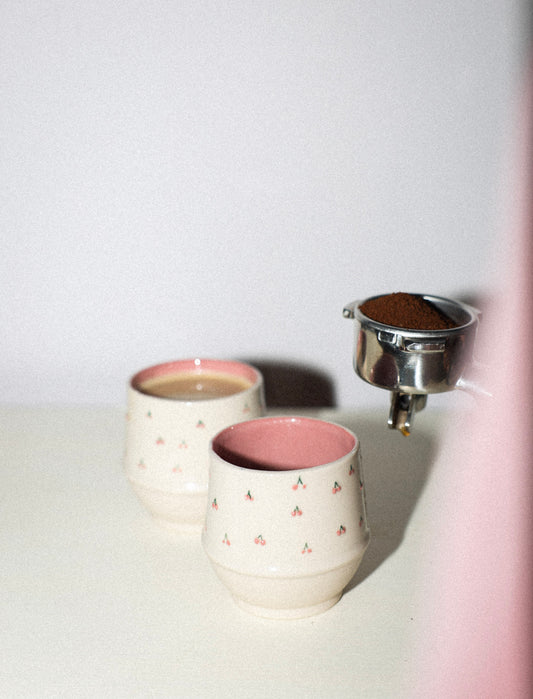 Two ceramic cups with pink interior and exterior, one being filled with a brown substance from a metal container.