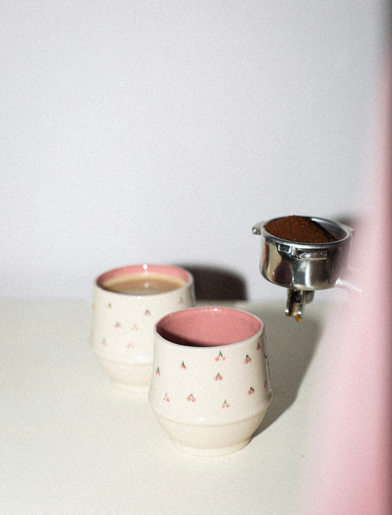 Two ceramic cups with pink interior and exterior, one being filled with a brown substance from a metal container.